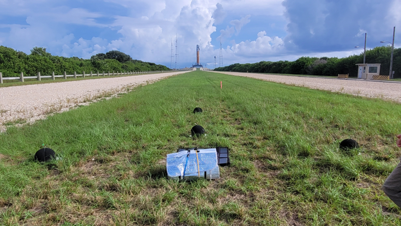 An array of four microphones (dark objects, two front, one left, one right) ready for noise measurements with NASA's Space Launch System in the background. (Credit: Kent Gee)